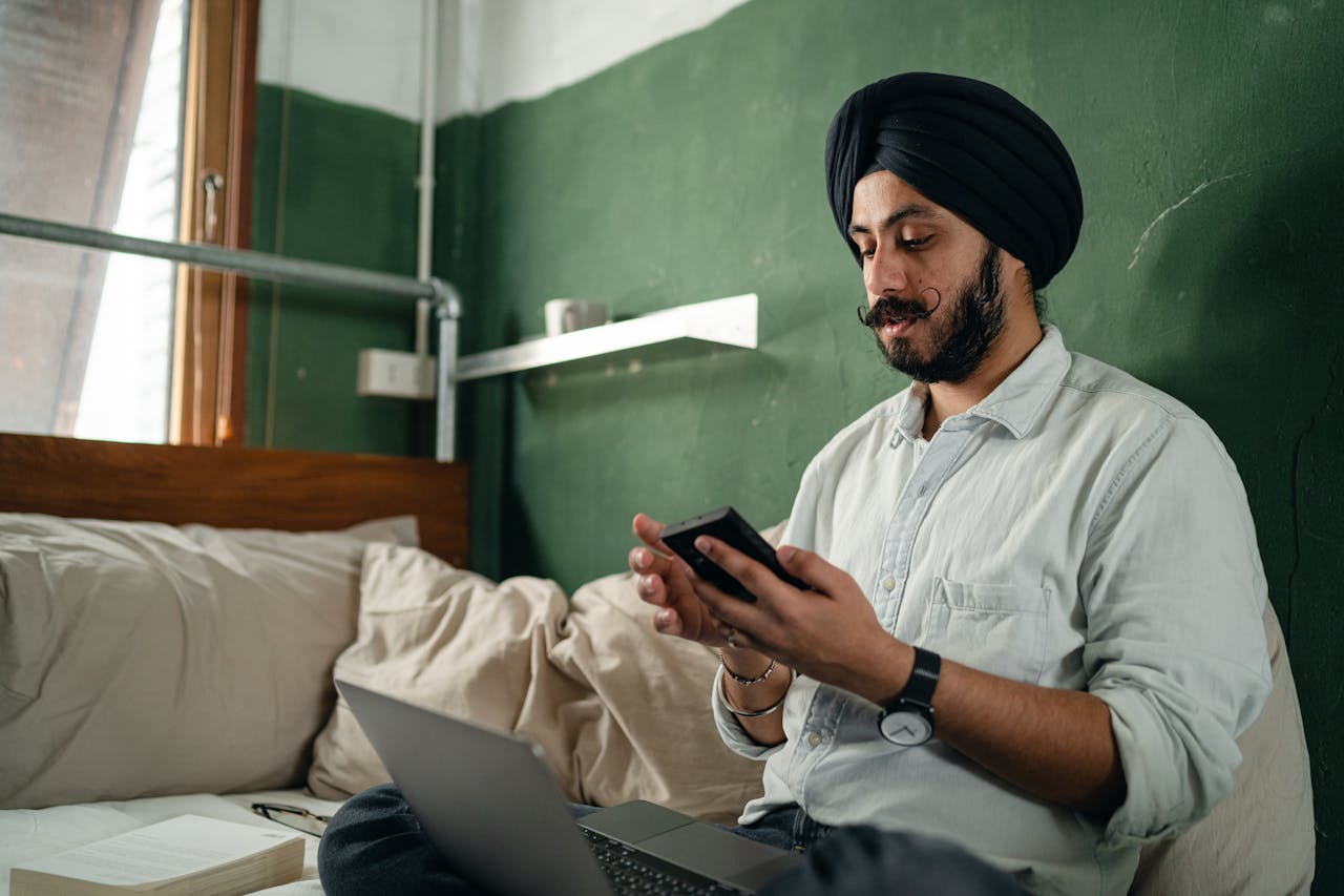 Concentrated bearded Sikh male wearing turban sitting on bed with pillows in green room near window with netbook and browsing smartphone pensively